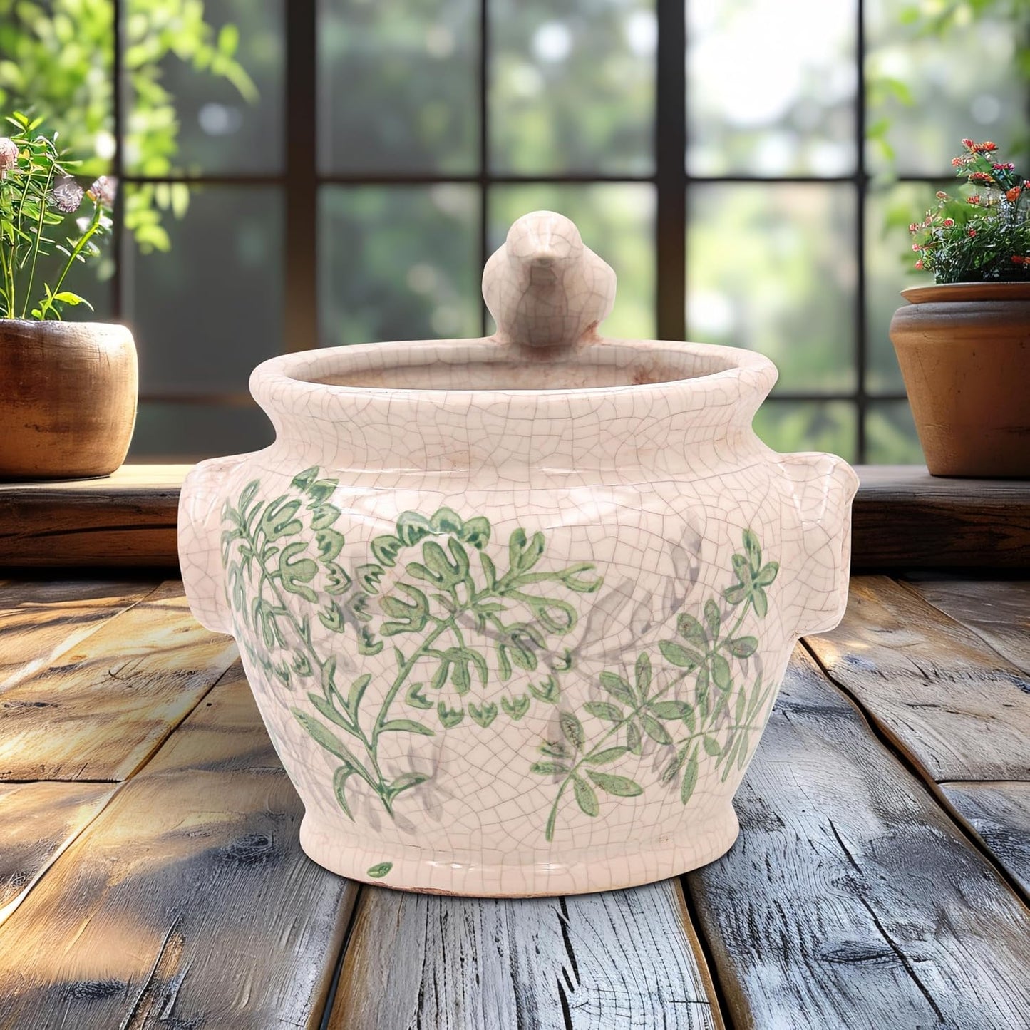 A ceramic lidded pot with a crackle glaze and green botanical pattern, displayed on a rustic wooden table in front of a window with potted plants.