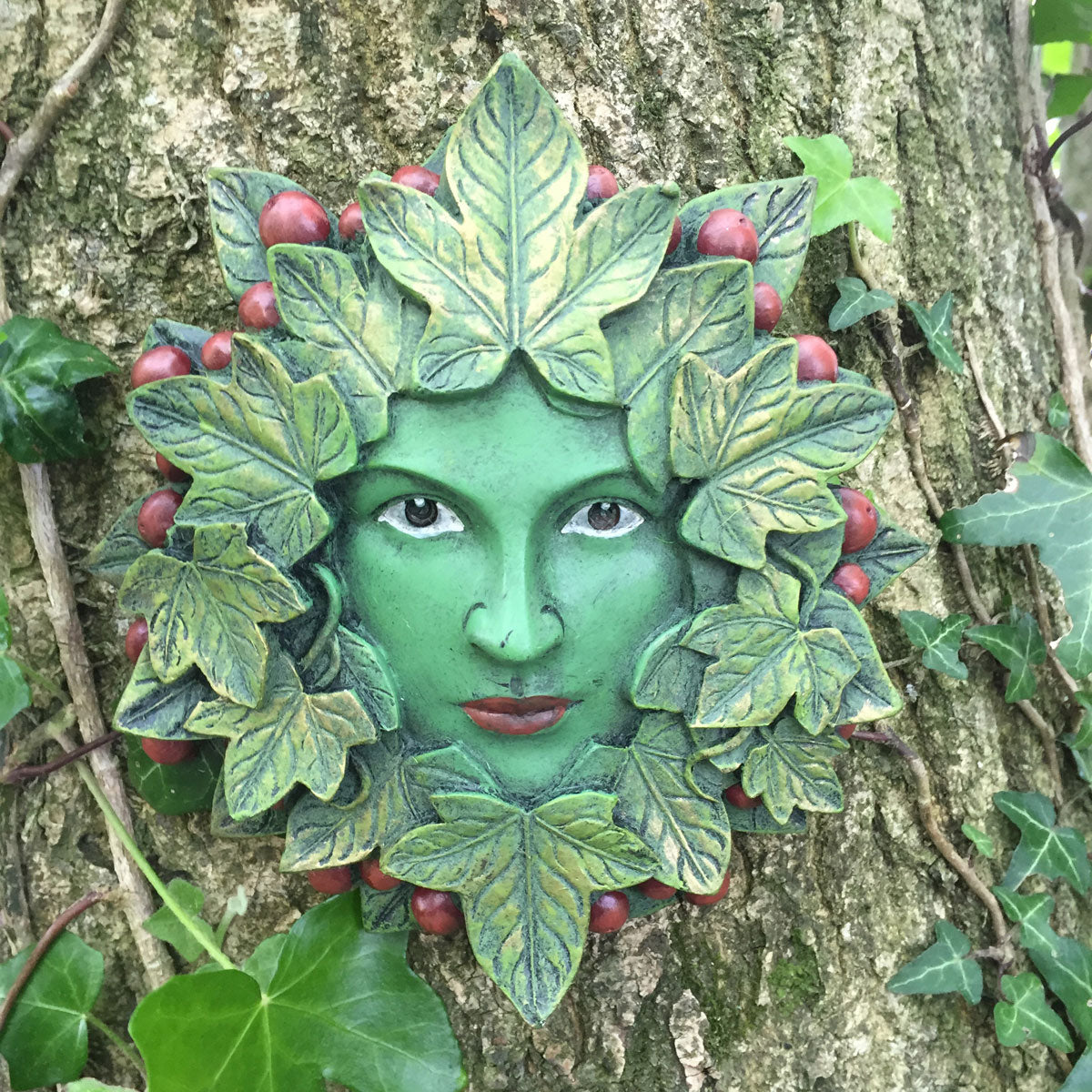 A green tree spirit wall plaque shaped like a face framed by leaves and berries, mounted on a tree trunk with ivy.