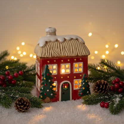 A small decorative gingerbread house ornament with a red facade, beige textured roof, white snow, green door and Christmas trees, and gold dot accents, shown on a plain white background.