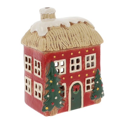 A small ceramic Christmas house ornament with a red body, beige thatched roof, white snow, green trees, and a green door, shown on a plain white background.