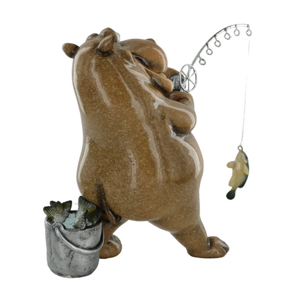 A ceramic figurine of a hippo holding a silver fishing rod with a fish, standing next to a small silver bucket containing two fish, on a light gray background.