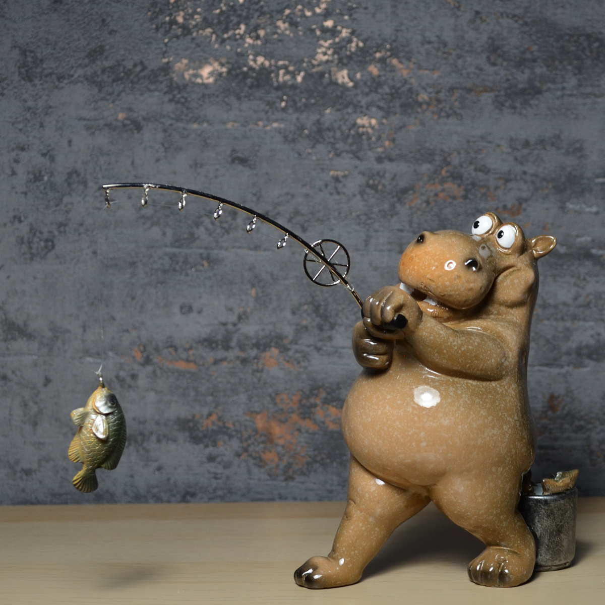 A ceramic hippo figurine holding a bent fishing rod with a small fish dangling from the line, set against a textured gray wall and light wood surface.