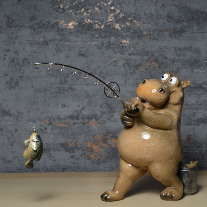 A ceramic hippo figurine holding a bent fishing rod with a small fish dangling from the line, set against a textured gray wall and light wood surface.