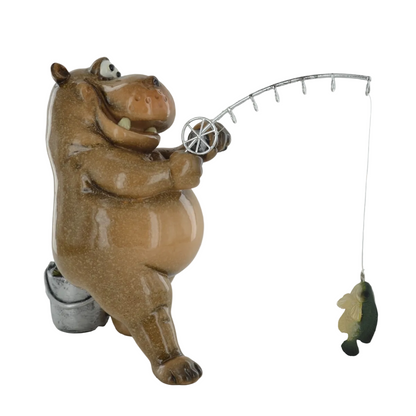 A ceramic hippopotamus figurine holding a silver fishing rod with a dangling fish, shown on a plain light gray background.