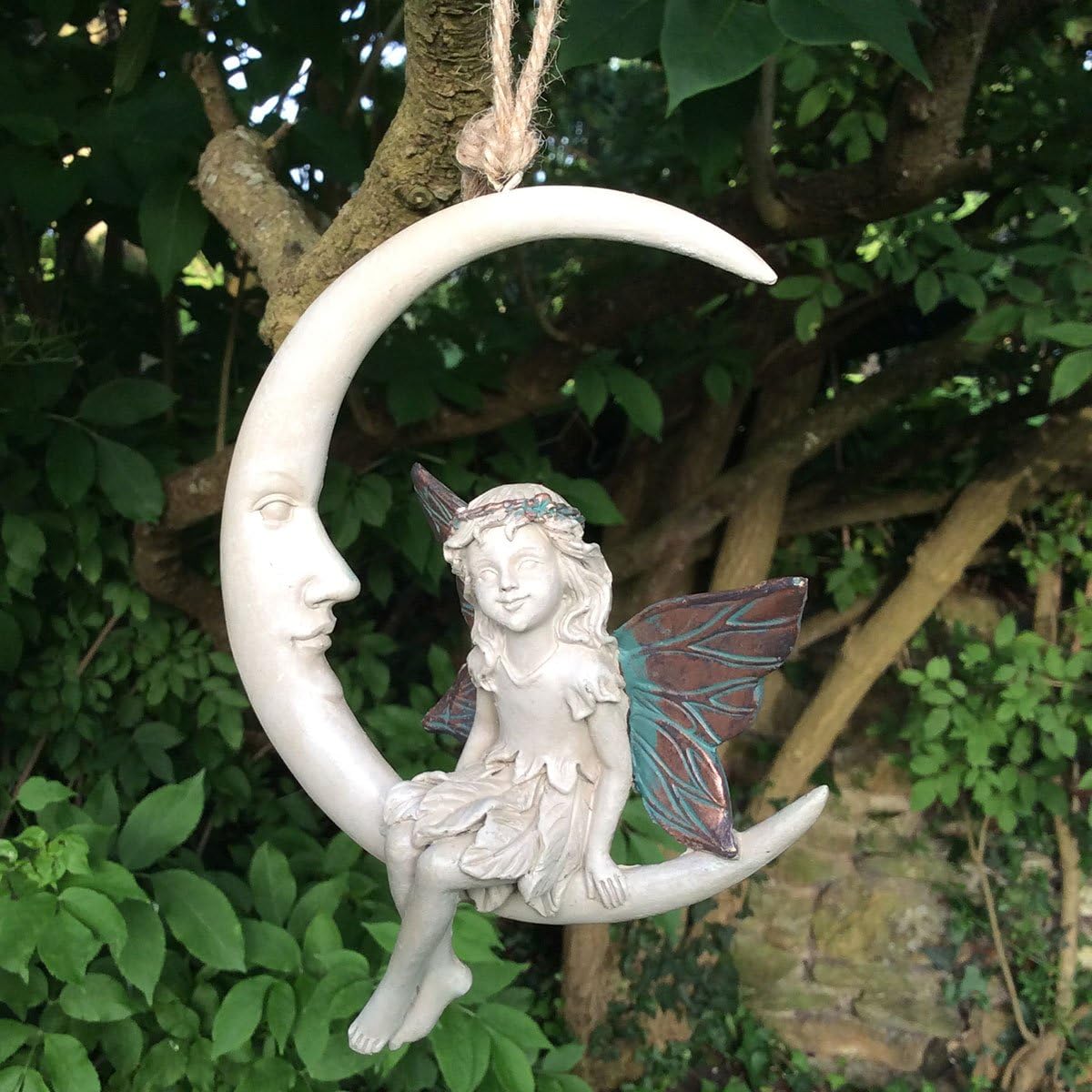 A decorative hanging ornament shaped like a crescent moon with a face and a fairy seated on it, photographed outdoors against a leafy tree background.