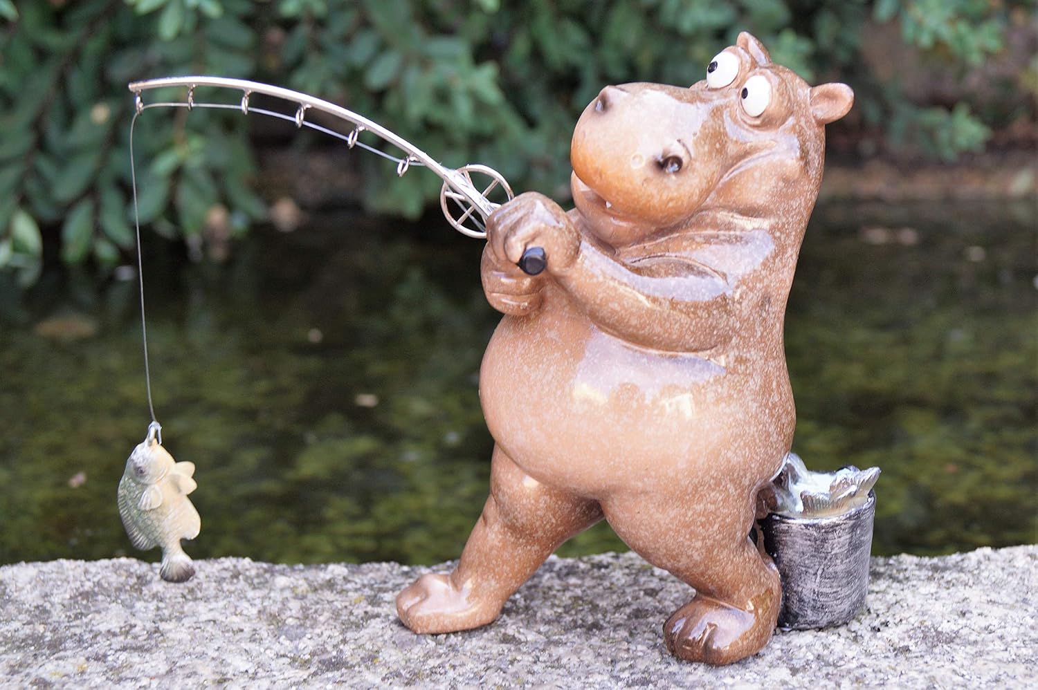 A ceramic hippo figurine holding a fishing rod with a fish dangling from the line, posed outdoors on a stone surface with water and greenery in the background.