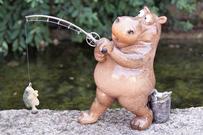 A ceramic hippo figurine holding a fishing rod with a fish dangling from the line, posed outdoors on a stone surface with water and greenery in the background.