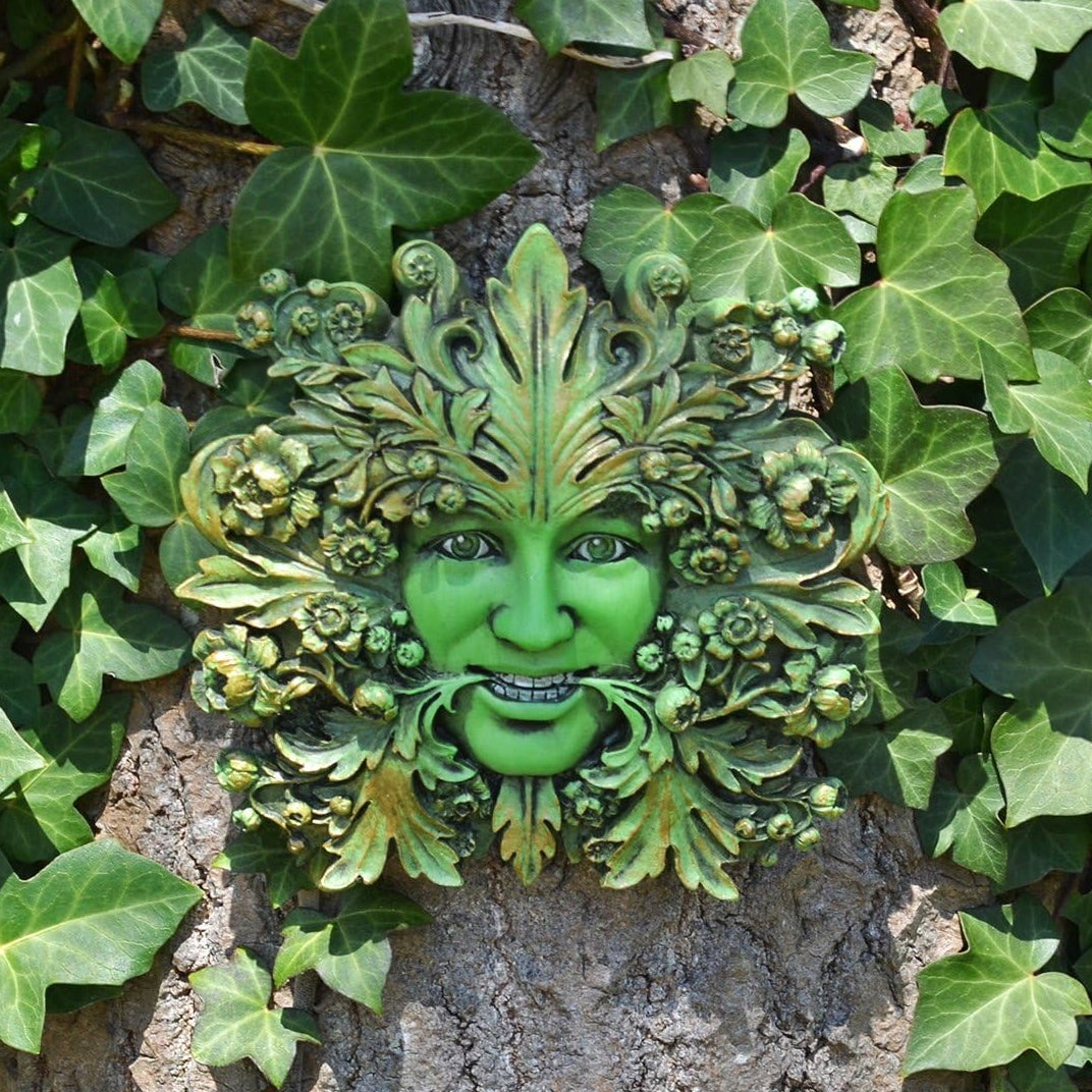A green garden wall plaque shaped like a smiling face surrounded by leaves and vines, mounted on a tree trunk and framed by real ivy outdoors.