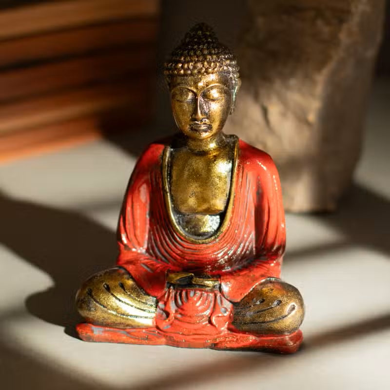 A small seated Buddha figurine with a gold-toned head, shoulders, and hands, and a red robe, photographed on a light surface with warm shadows and a blurred stone-like object in the background.