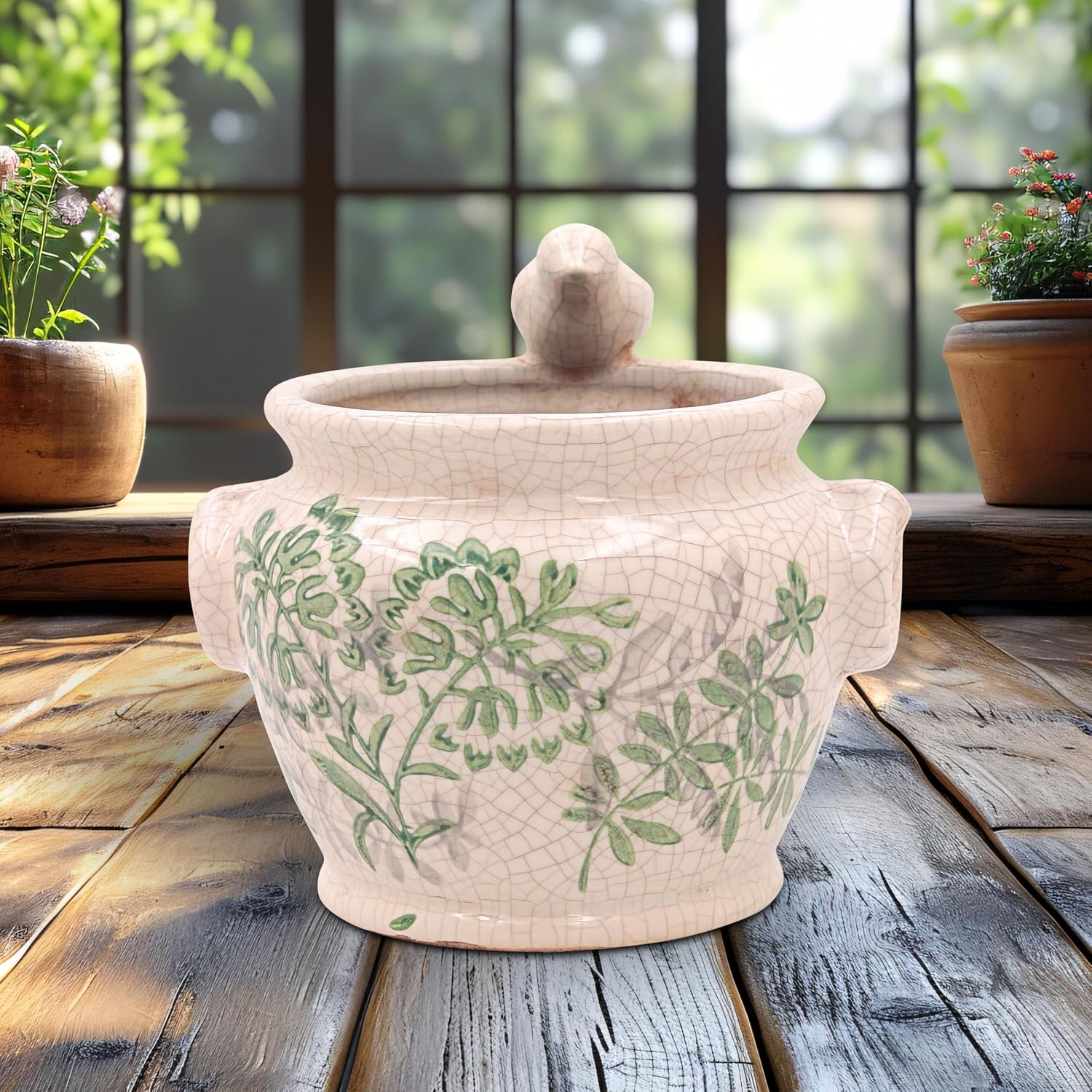 A ceramic lidded pot with a crackle glaze and green botanical pattern, displayed on a rustic wooden table in front of a window with potted plants.