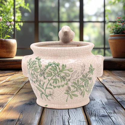 A ceramic lidded pot with a crackle glaze and green botanical pattern, displayed on a rustic wooden table in front of a window with potted plants.