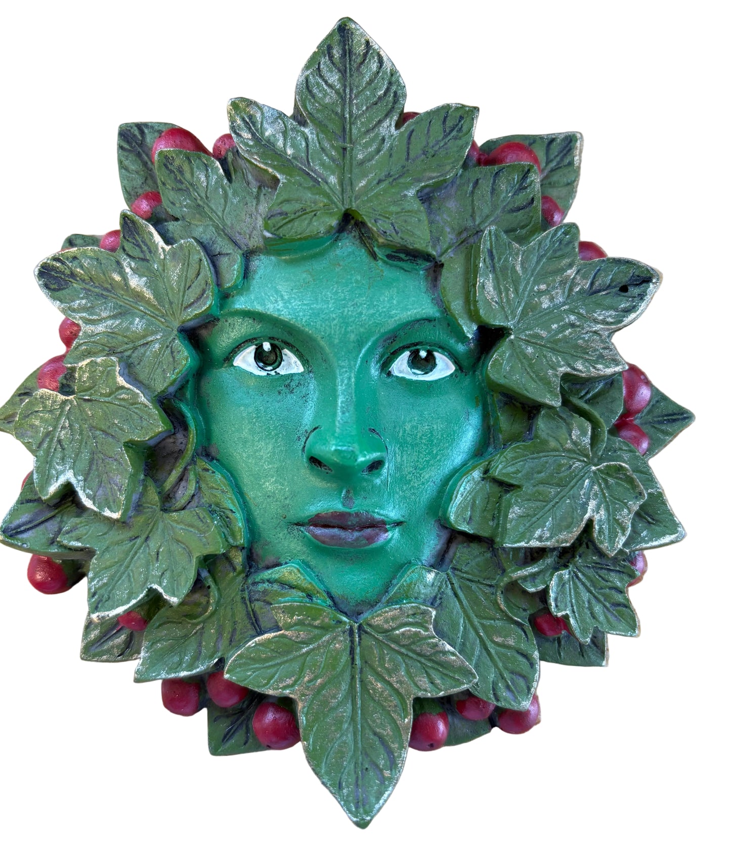 A green face mask or decorative figurine with leafy vines and small red berries, photographed from above on a light wood surface.