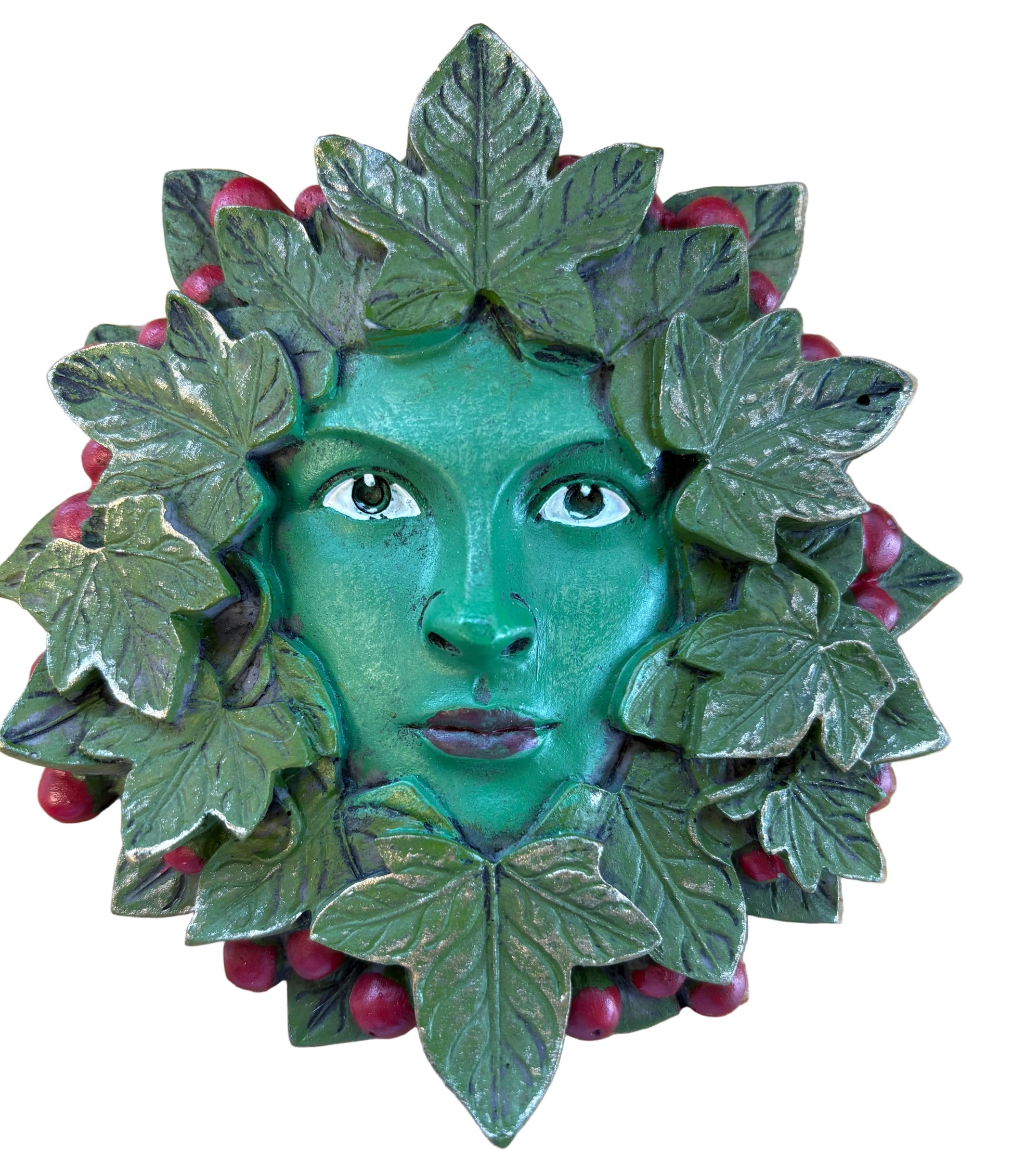 A green face mask or decorative figurine with leafy vines and small red berries, photographed from above on a light wood surface.
