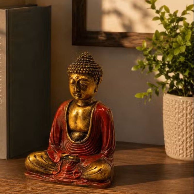 A small seated Buddha figurine with a gold-toned head and torso and red draped robes, displayed on a wooden shelf beside books and a potted plant.